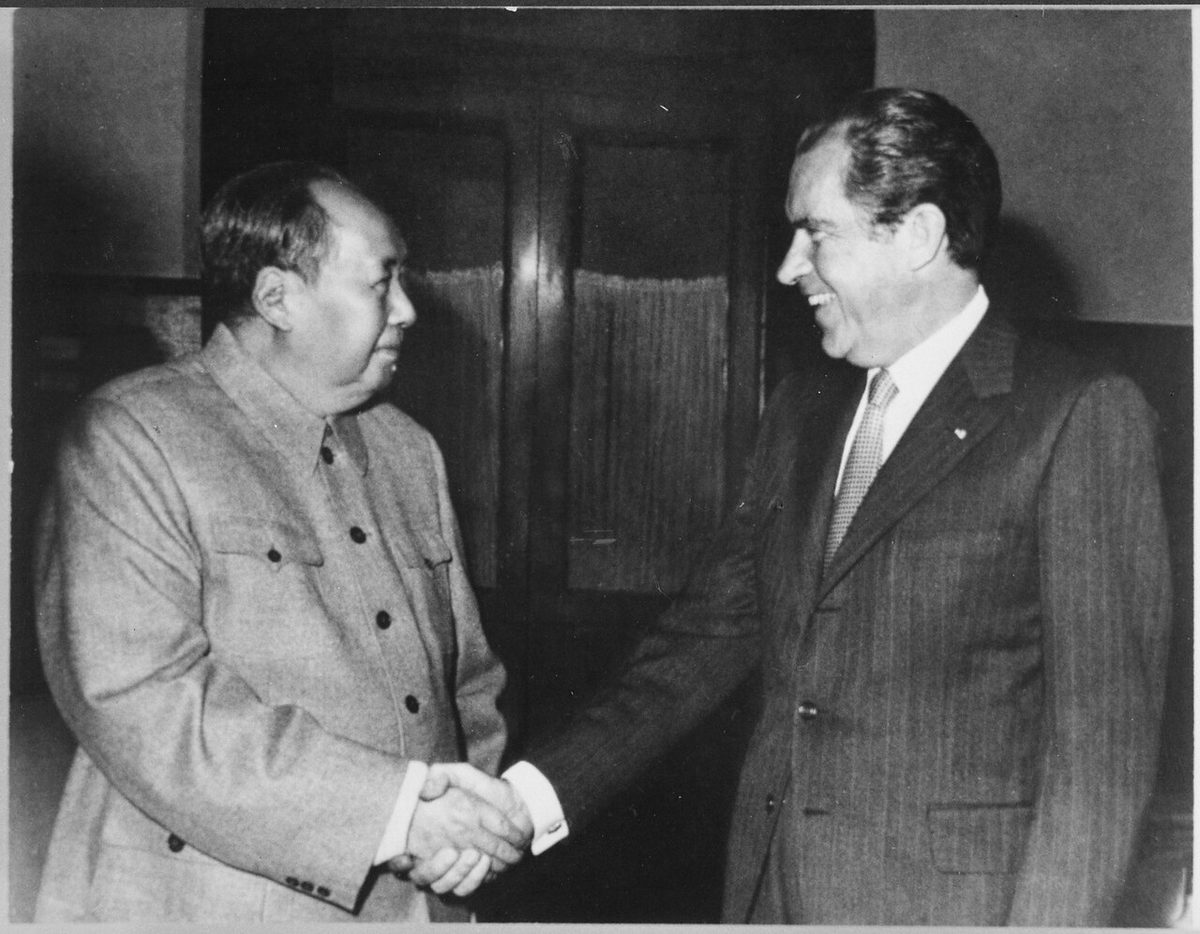 Mao Zedong, sentado, aperta a mão de Richard Nixon numa sala com poltronas e estantes de livros.
